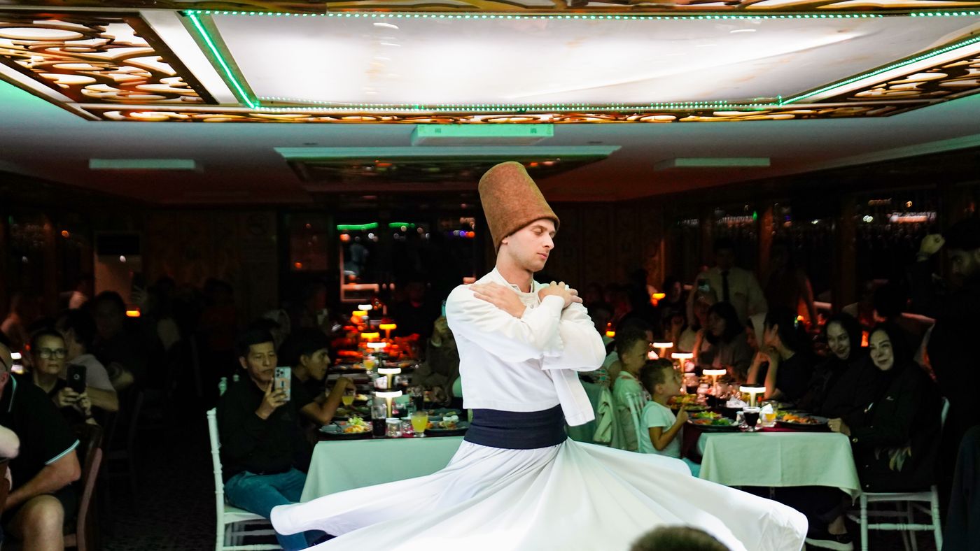 The dervish turns between tables in the lamplit salon