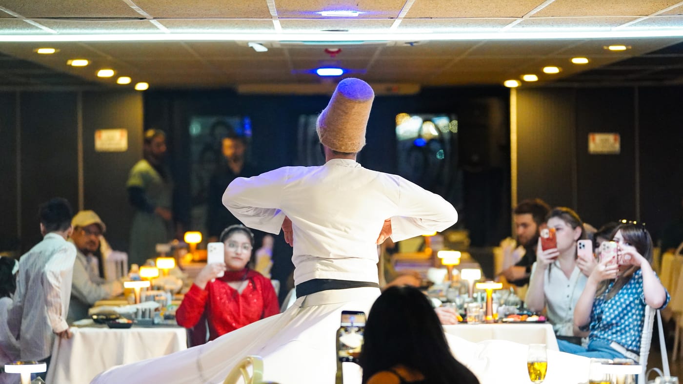 The dervish turns between tables in the lamplit salon